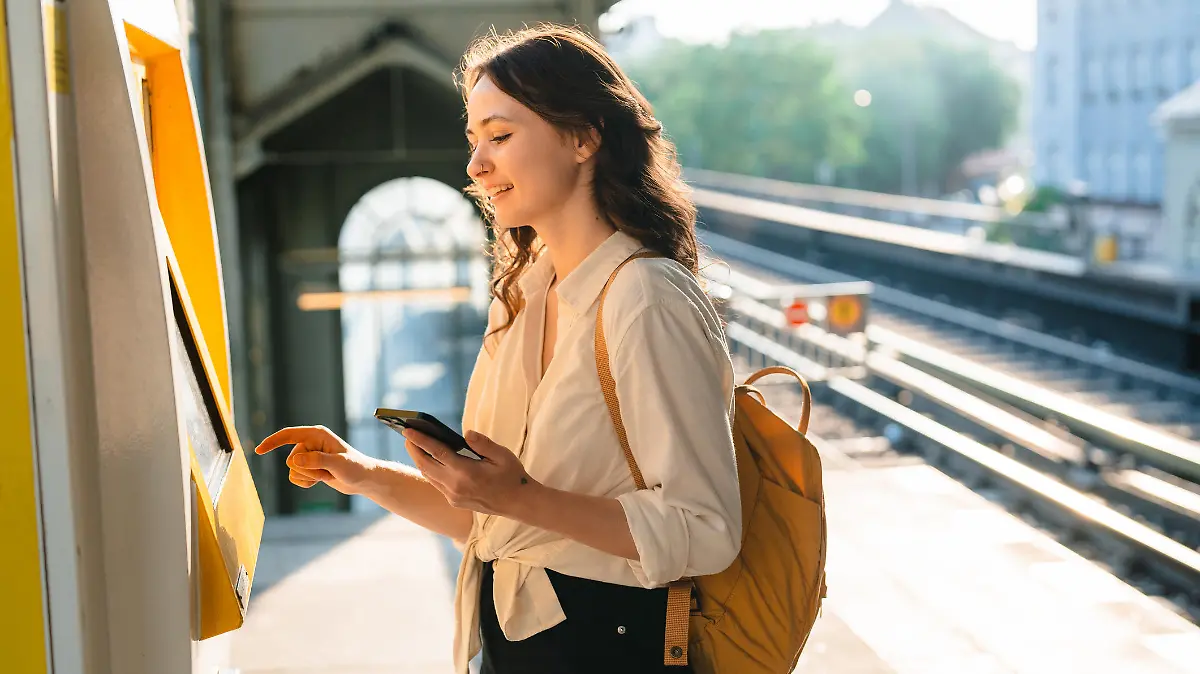 Cheerful woman  buying ticket from ticket machine on the station while holding smartphone