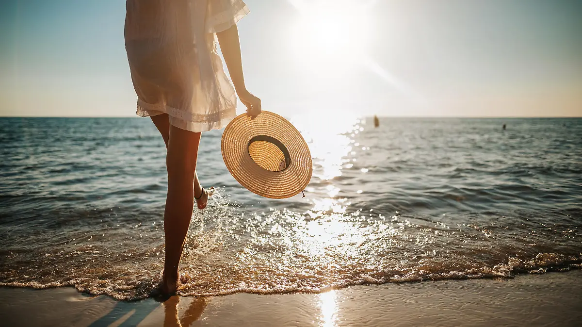 Close-up of young woman in white sun dress and with hat in hand walking alone on sandy beach at summer sunset, splashing water in sea shallow