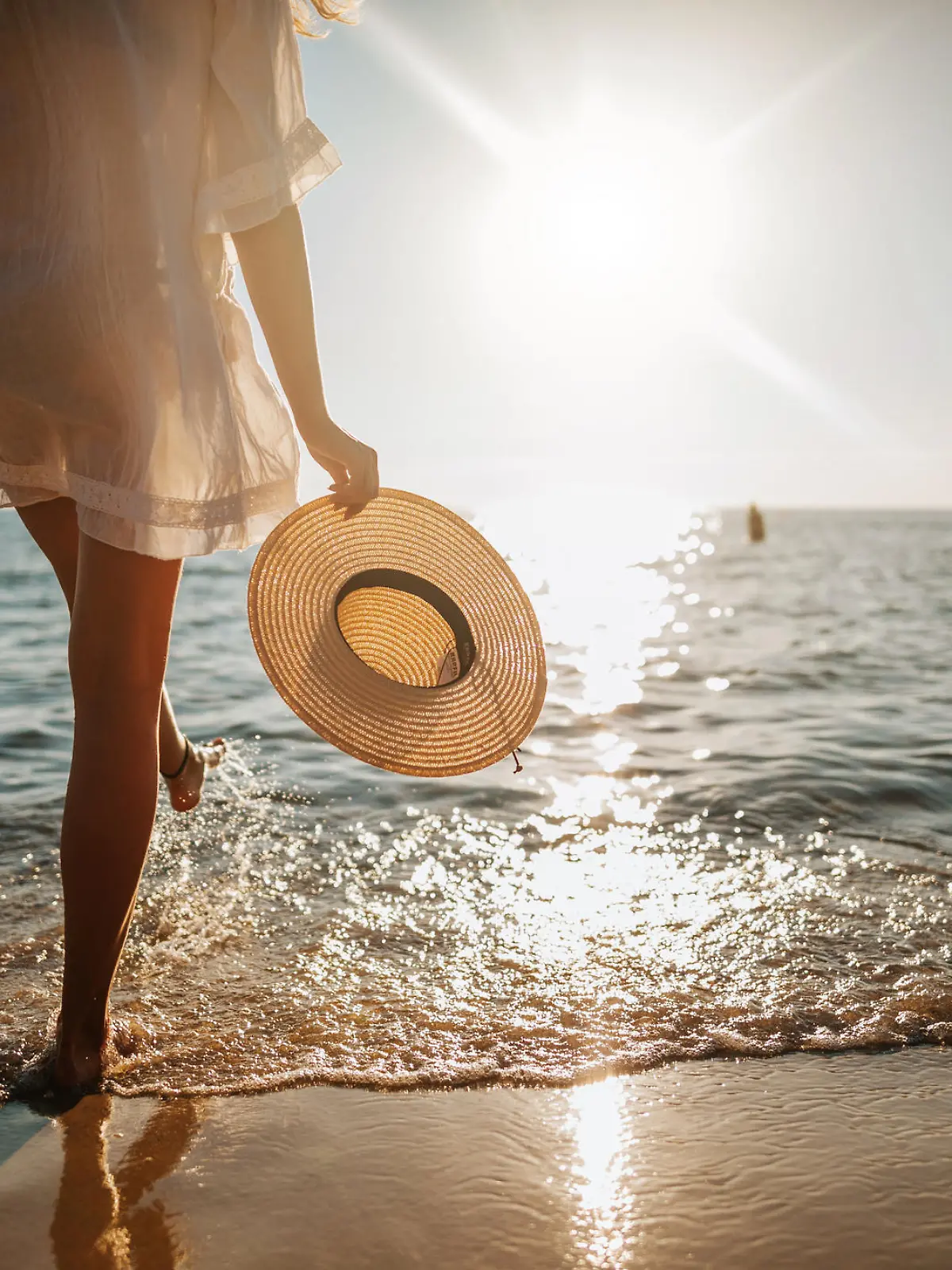 Close-up of young woman in white sun dress and with hat in hand walking alone on sandy beach at summer sunset, splashing water in sea shallow