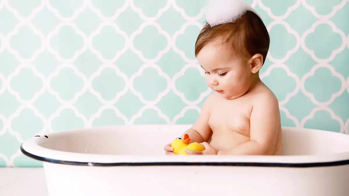 A baby looks at a rubber ducky as she sits in an antique bathtub. Copy space in vintage wallpaper. 