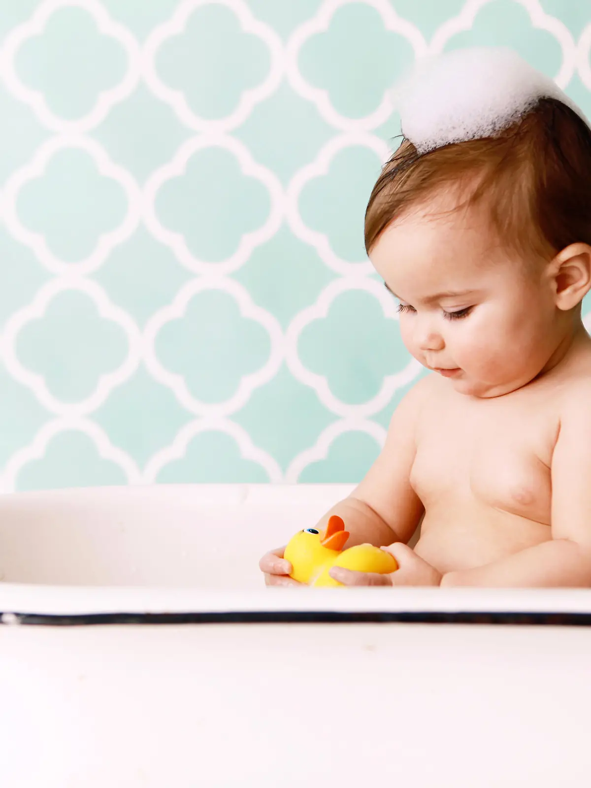 A baby looks at a rubber ducky as she sits in an antique bathtub. Copy space in vintage wallpaper. 