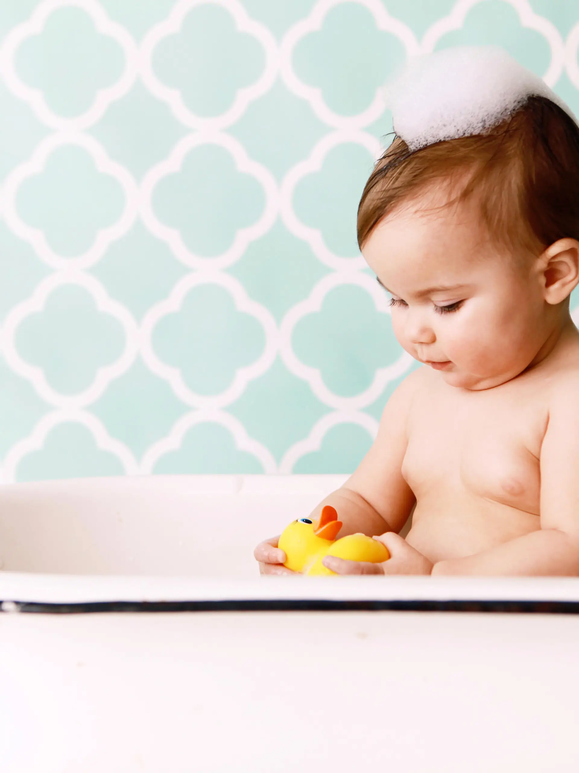 A baby looks at a rubber ducky as she sits in an antique bathtub. Copy space in vintage wallpaper. 