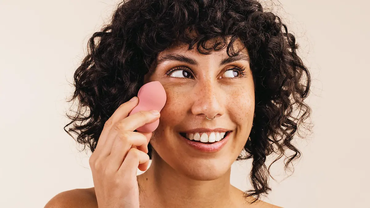 Pretty young woman looking away with a smile while using a makeup sponge on her face. Happy young woman using a beauty blender to apply makeup foundation on her face.