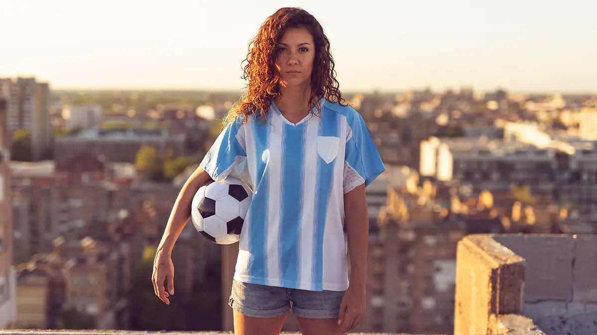 Young woman wearing a football jersey standing on a building rooftop, holding a ball and watching a sunset over the city.