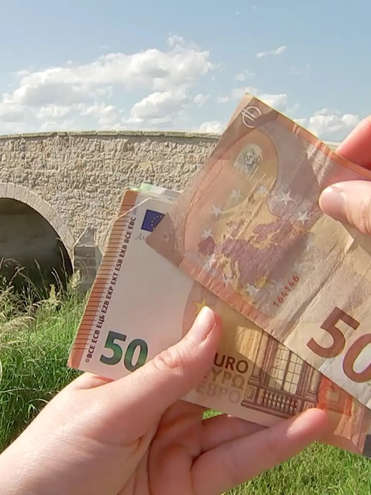 Die Brücke mit Zaun im Hintergrund, davor hält eine Hand Geldscheine.