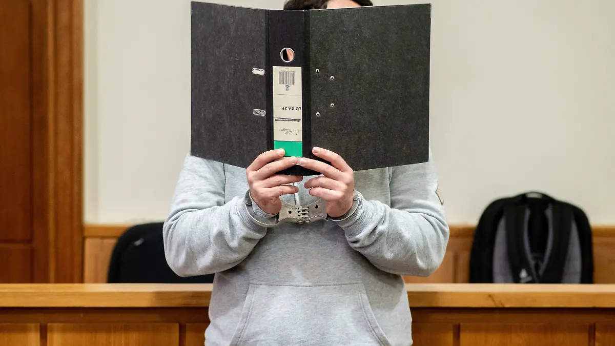 Ein Angeklagter steht mit Handfesseln in einem Saal im Landgericht Oldenburg und verdeckt sein Gesicht mit einem Aktenordner. Dem Angeklagten wird die Anstiftung zu einem versuchten Mord vorgeworfen, einem weiteren Angeklagten die Ausführung des versuchten Mordes in Tateinheit mit gefährlicher Körperverletzung. Der Mann soll aus Eifersucht beschlossen haben, seine Ex-Frau durch eine dritte Person töten zu lassen.