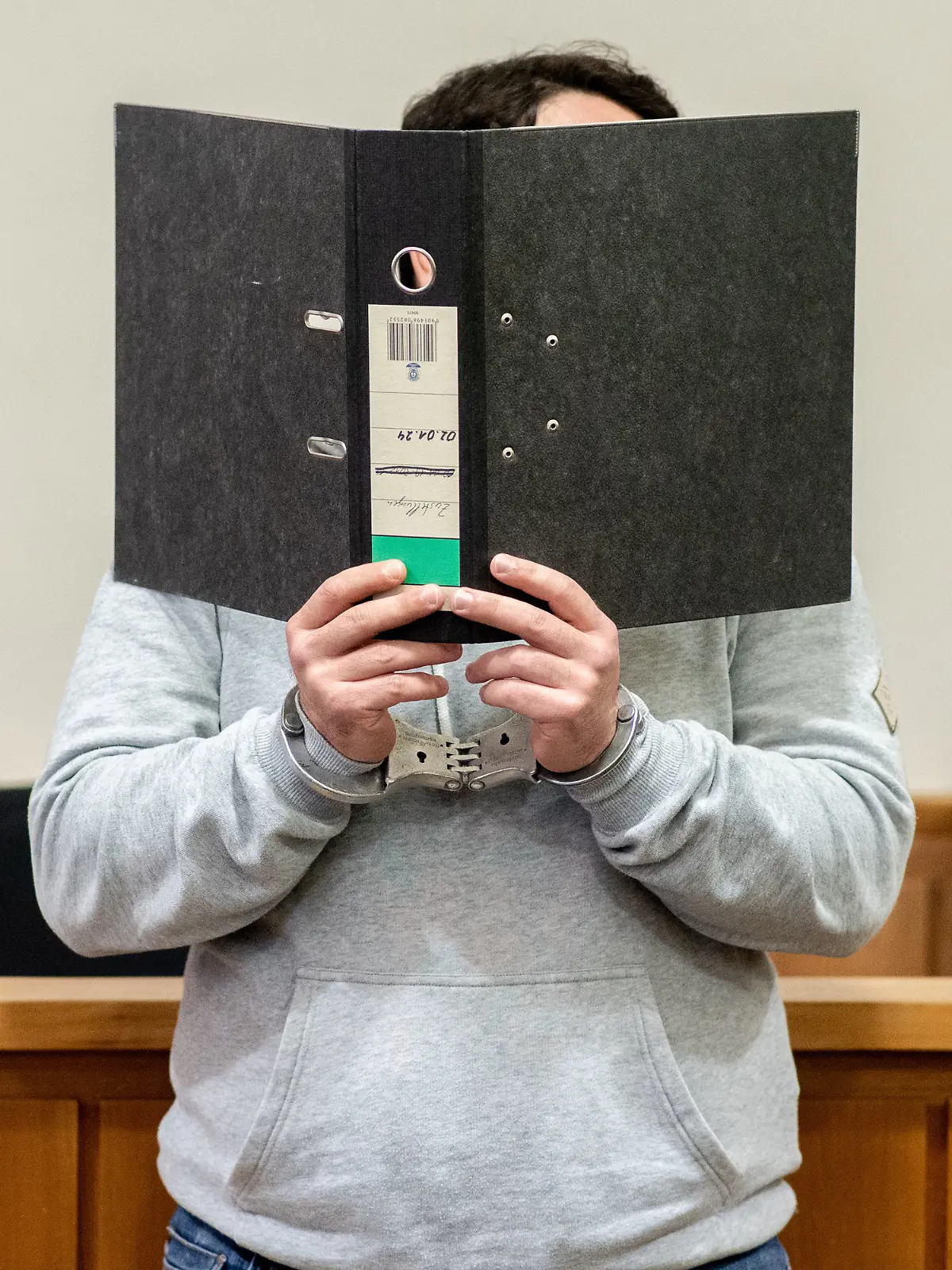 Ein Angeklagter steht mit Handfesseln in einem Saal im Landgericht Oldenburg und verdeckt sein Gesicht mit einem Aktenordner. Dem Angeklagten wird die Anstiftung zu einem versuchten Mord vorgeworfen, einem weiteren Angeklagten die Ausführung des versuchten Mordes in Tateinheit mit gefährlicher Körperverletzung. Der Mann soll aus Eifersucht beschlossen haben, seine Ex-Frau durch eine dritte Person töten zu lassen.