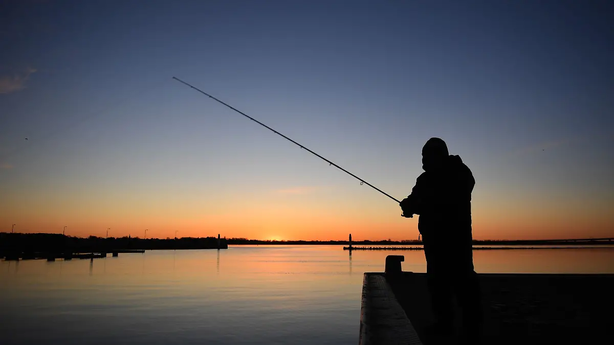 Die aufgehende Sonne vor einem Angler taucht den Himmel in ein warmes Licht.