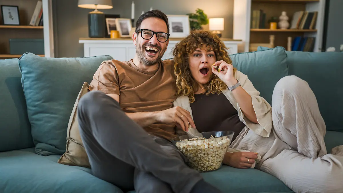 Couple man and woman caucasian husband and wife or boyfriend and girlfriend sit on the sofa bed at home watch tv movie series with bowl of popcorn happy smile bonding love family concept copy space