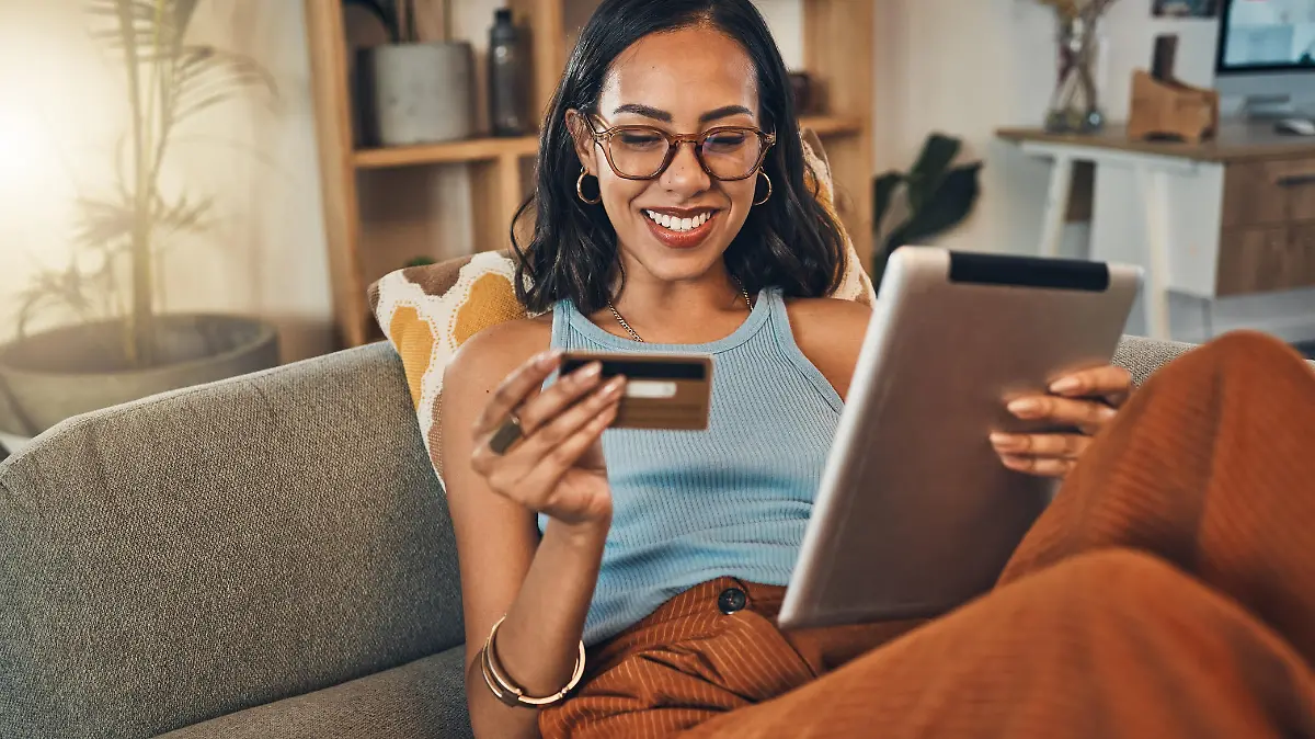 Smiling mixed race woman using credit card for ecommerce on digital tablet at home. Happy hispanic sitting alone on living room sofa, using technology for ebanking. Relaxing, ordering, buying online