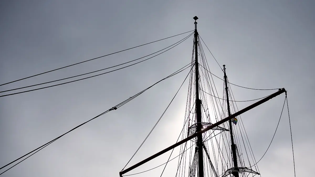Die Masten eines Segelschiffes zeichnen sich bei trübem Wetter vor dem bewölktem Himmel ab.