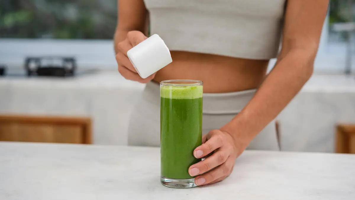 An unrecognizable woman is adding a protein powder in a glass of green smoothie.