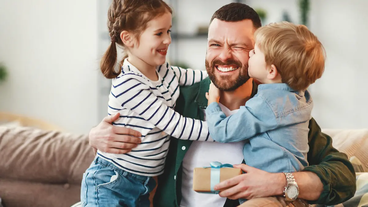 Joyful man with gift box in hand sitting on sofa at home and hugging cheerful son and daughter giving congratulations on fathers day