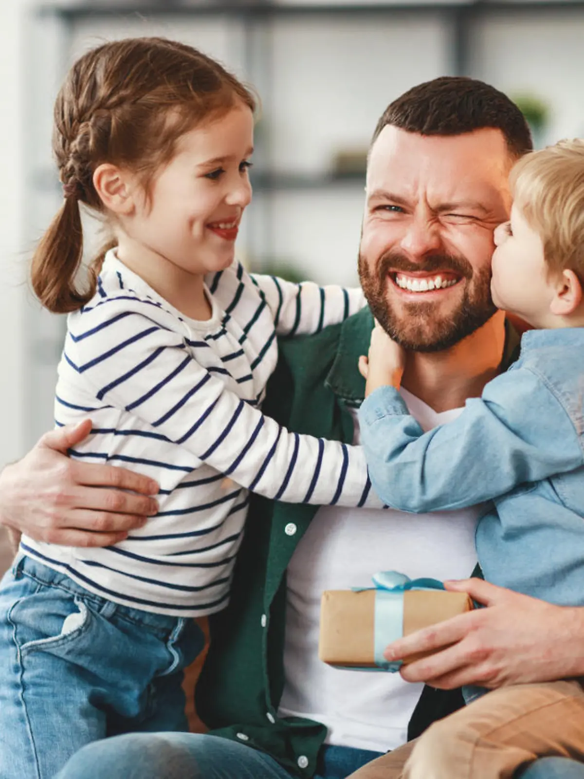 Joyful man with gift box in hand sitting on sofa at home and hugging cheerful son and daughter giving congratulations on fathers day