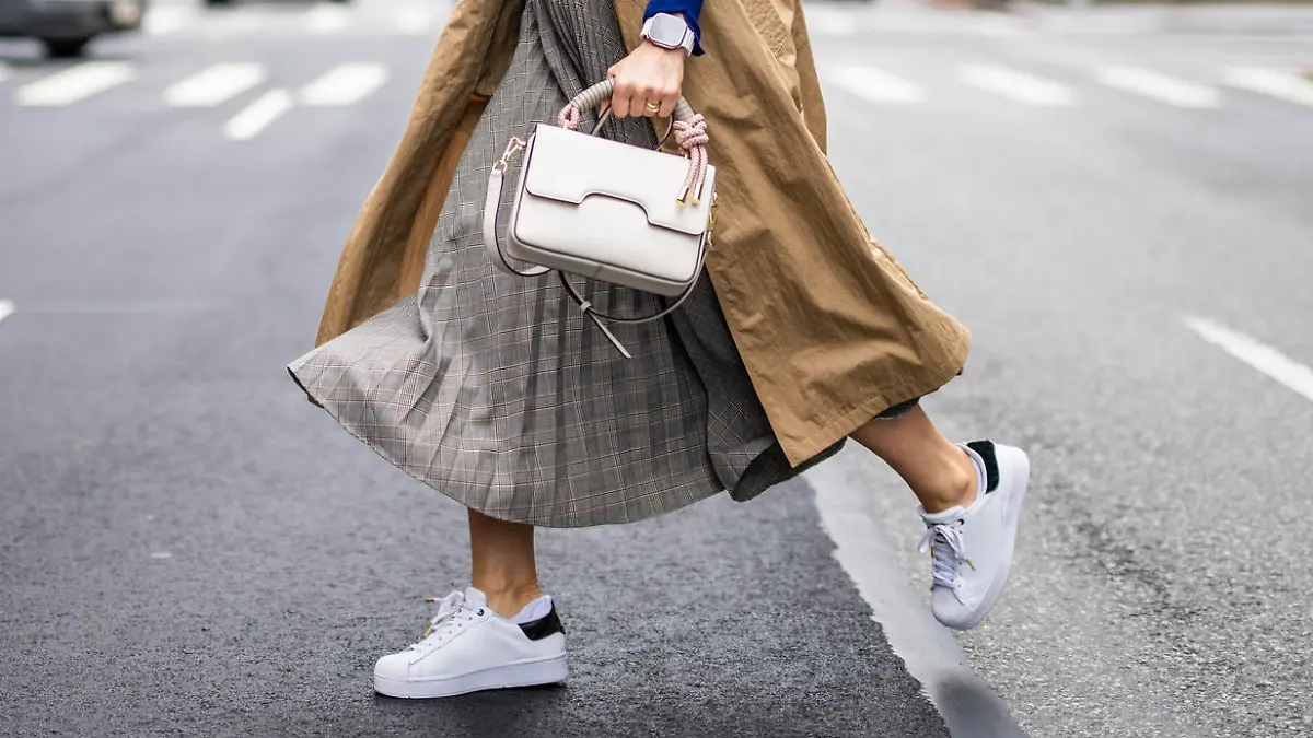 Beautiful woman in a beige sleeveless coat, skirt, turtleneck sweater, white sneakers and with a handbag seen crossing a street in Manhattan, New York, while using her wireless headphones and talking on the phone during a break at work.