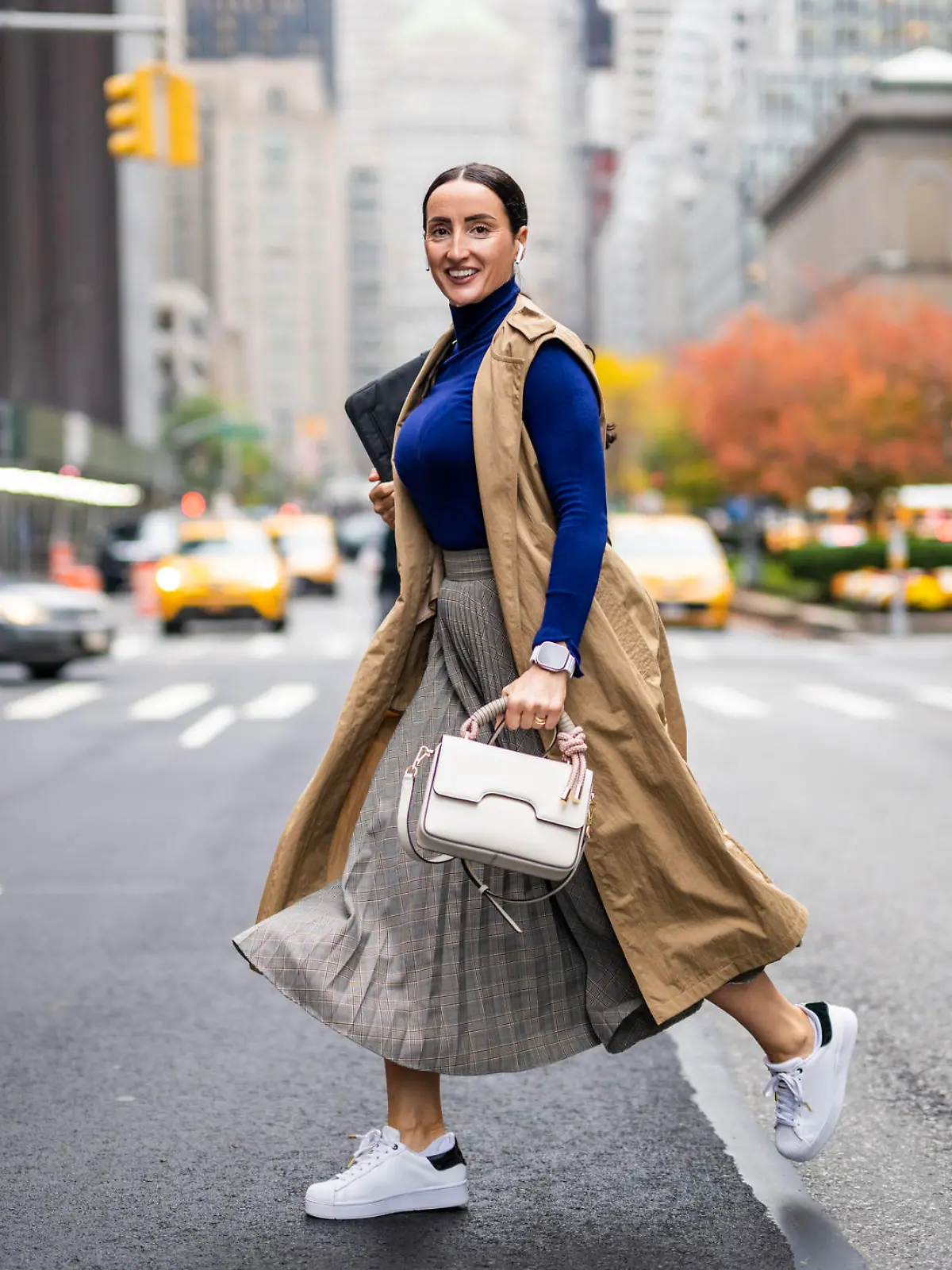 Beautiful woman in a beige sleeveless coat, skirt, turtleneck sweater, white sneakers and with a handbag seen crossing a street in Manhattan, New York, while using her wireless headphones and talking on the phone during a break at work.