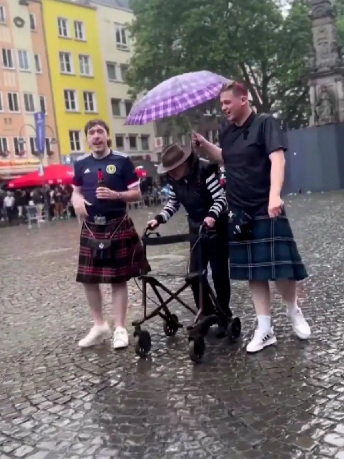 Zwei Schotten halten einer älteren Dame in Köln den Regenschirm, während sie mit ihrem Rollator läuft.