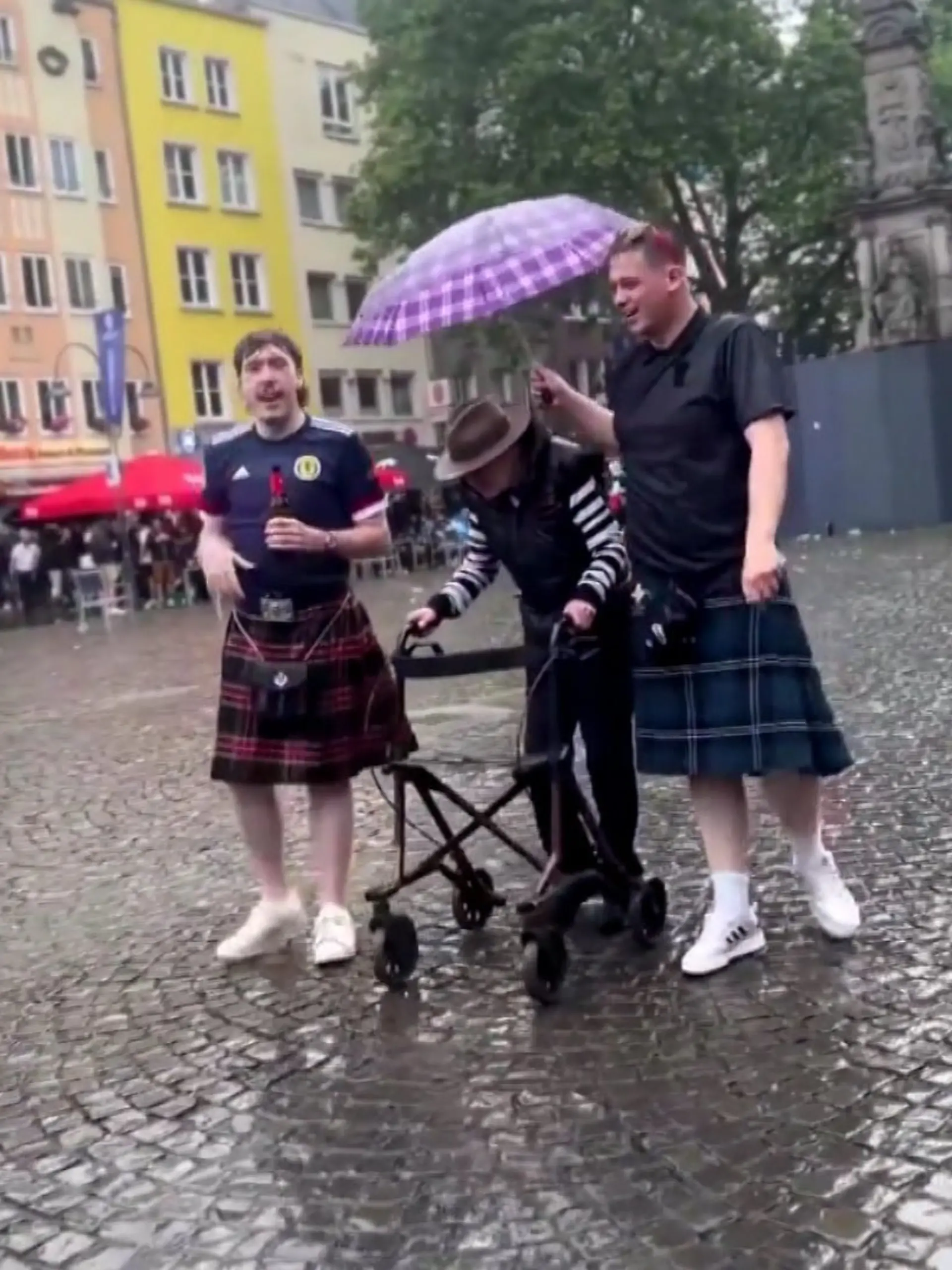 Zwei Schotten halten einer älteren Dame in Köln den Regenschirm, während sie mit ihrem Rollator läuft.