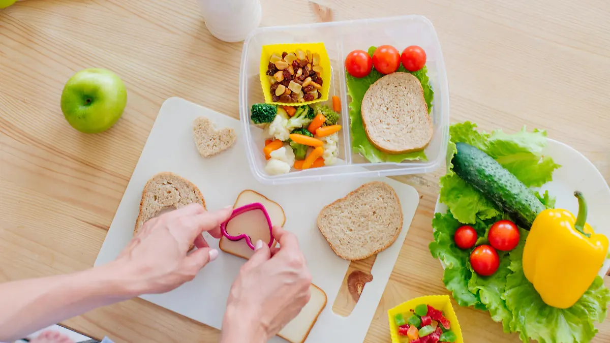 Healthy food concept. Closeup of female hands preparing sandwich for lunch box filled with bread, fresh vegetables, fruits and snack on the wooden table. Top view, selective focus.