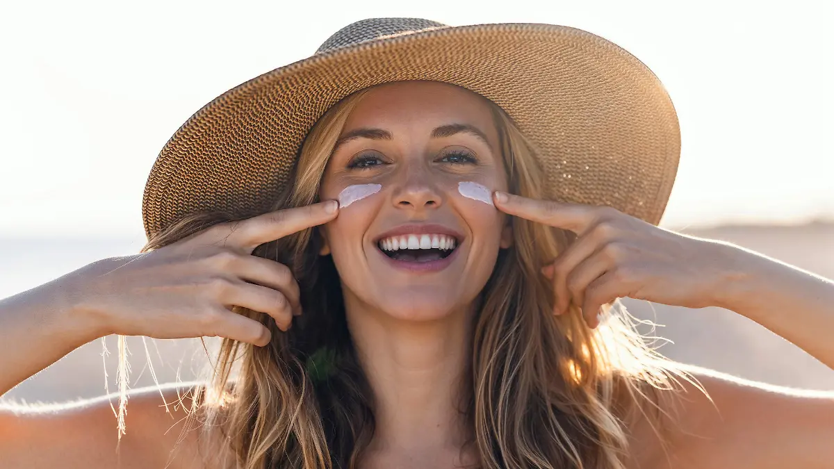 Portrait of beautiful smiling woman applying sunscreen on her face while looking at camera at the beach.