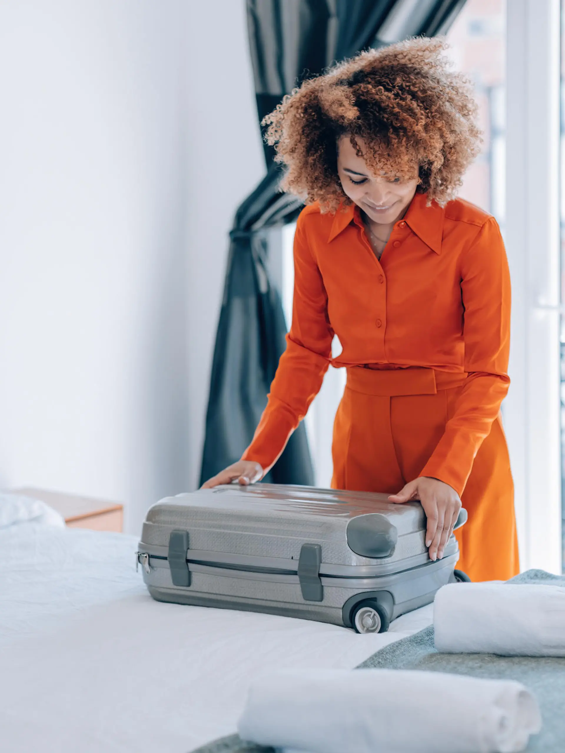 curly hair brazilian elegant young woman in orange suit open her luggage in hotel room bed