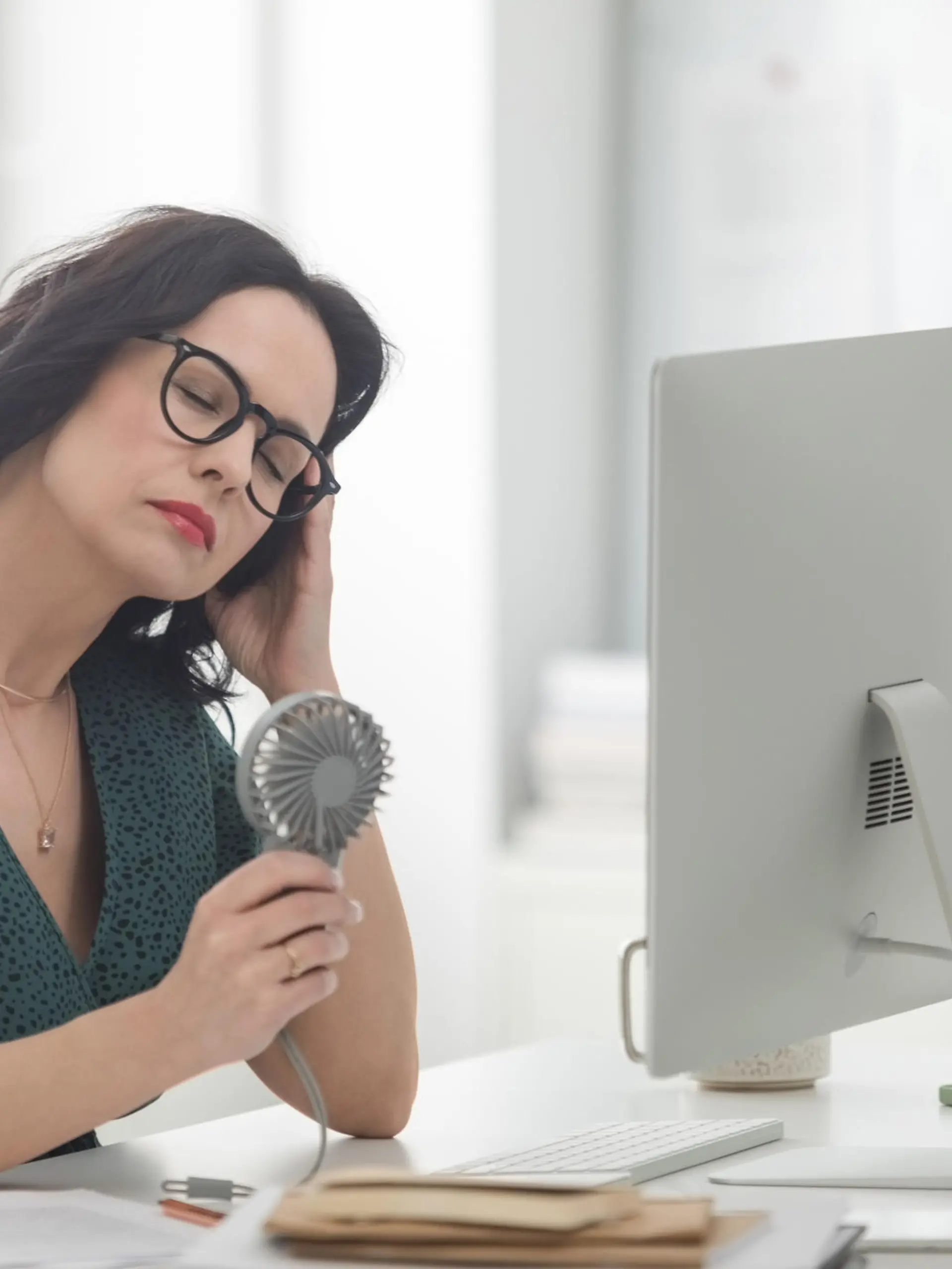 Menopausal mature woman wearing green dress sitting at the desk in the office in front of computer, having hot flashes and using hand fan.
