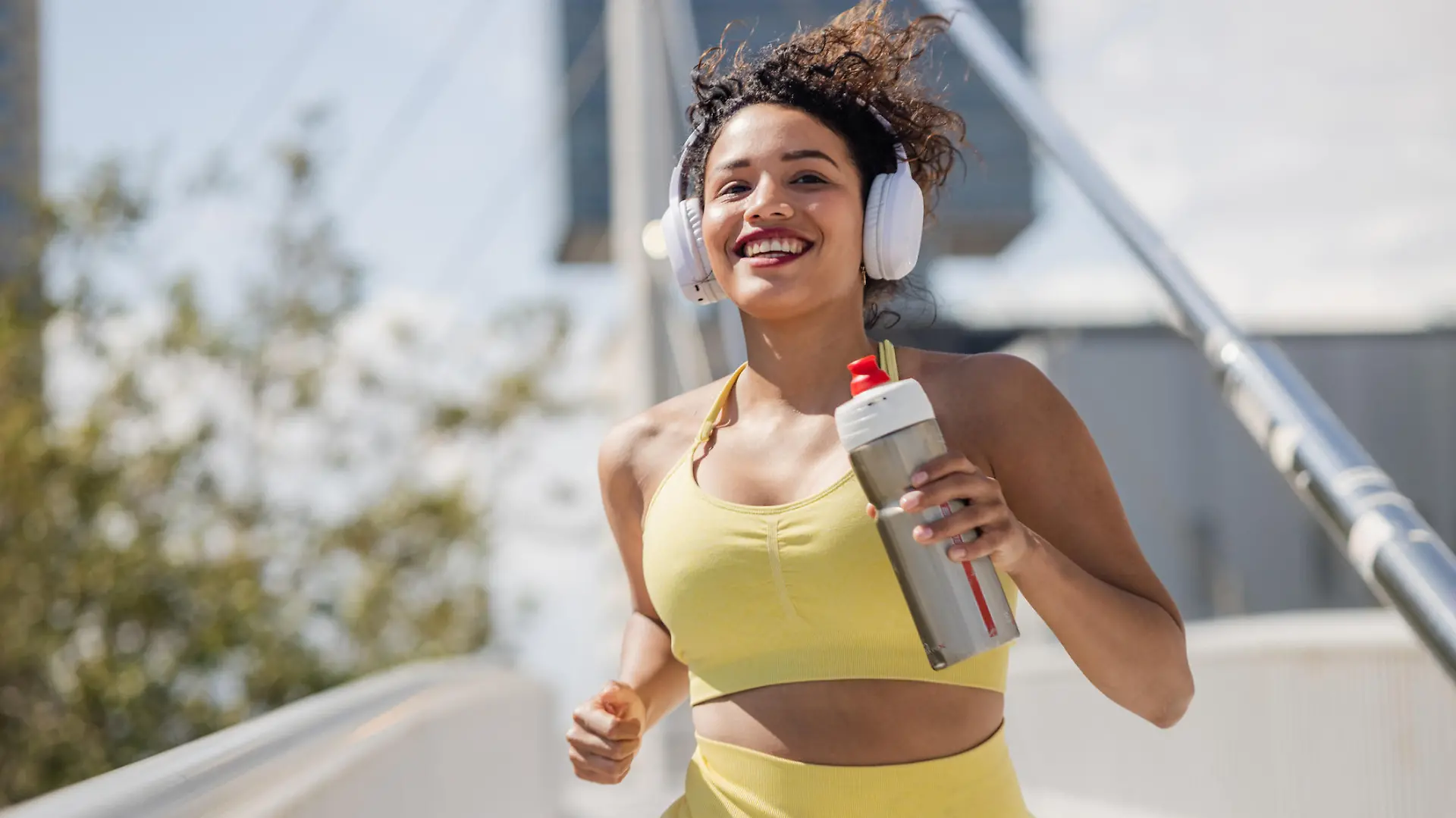 Portrait of a cheerful woman with wireless headphones running on a sunny day. She is holding a water bottle while running.