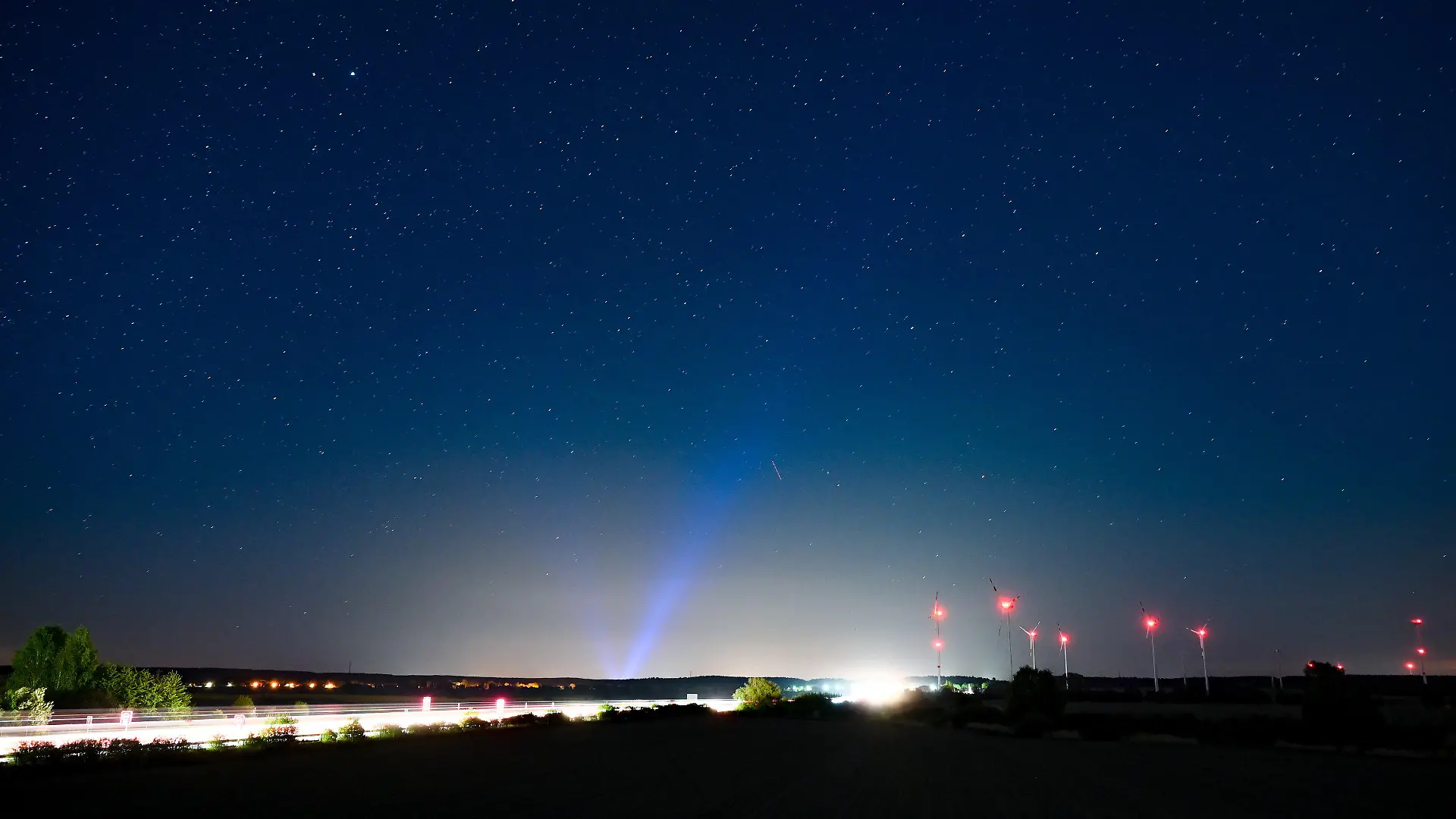 PRODUKTION - 06.05.2022, Brandenburg, Jacobsdorf: Hell erleuchtet ist die Autobahn A12, am Rand das rote Licht der Windenergieanlagen und im Hintergrund erhellt blaues Licht den Nachthimmel über der Stadt Frankfurt (Oder). Angestrahlte Kirchen und Denkmäler, Flutlicht im Stadien und grelle Straßenbeleuchtung - die Menschen machen vielerorts die Nacht zum Tag, mit verheerenden Folgen für die Tierwelt und die Artenvielfalt. (zu dpa-KORR.: "Schattenseite des Lichts: Lichtverschmutzung wird für Tiere zur Qual") Foto: Patrick Pleul/dpa +++ dpa-Bildfunk +++