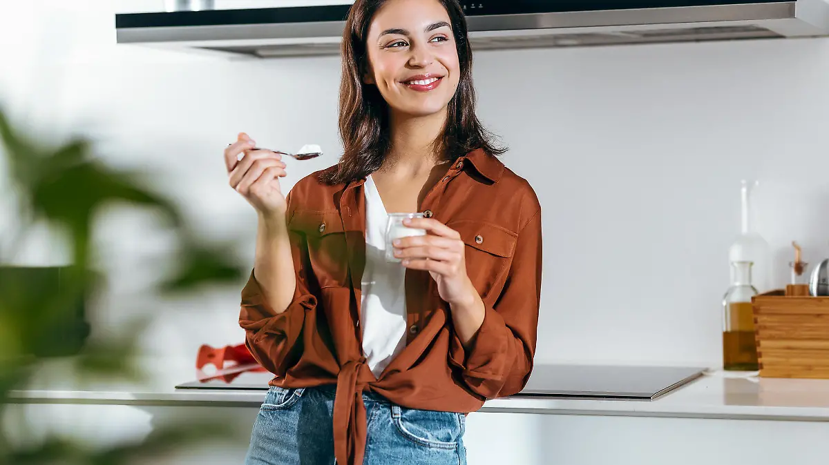 Shot of beautiful happy woman eating a yoghurt while looking forwards in the kitchen at home
