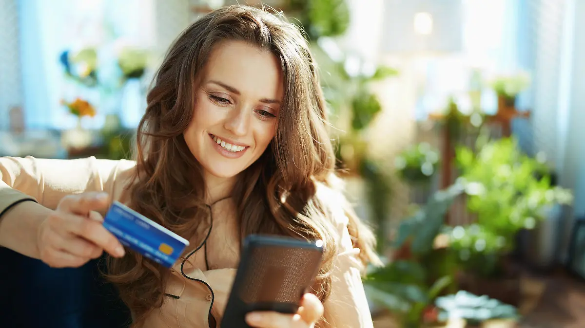 Green Home. happy trendy middle aged woman with long wavy hair with credit card making online shopping on e-commerce website in the modern house in sunny day.