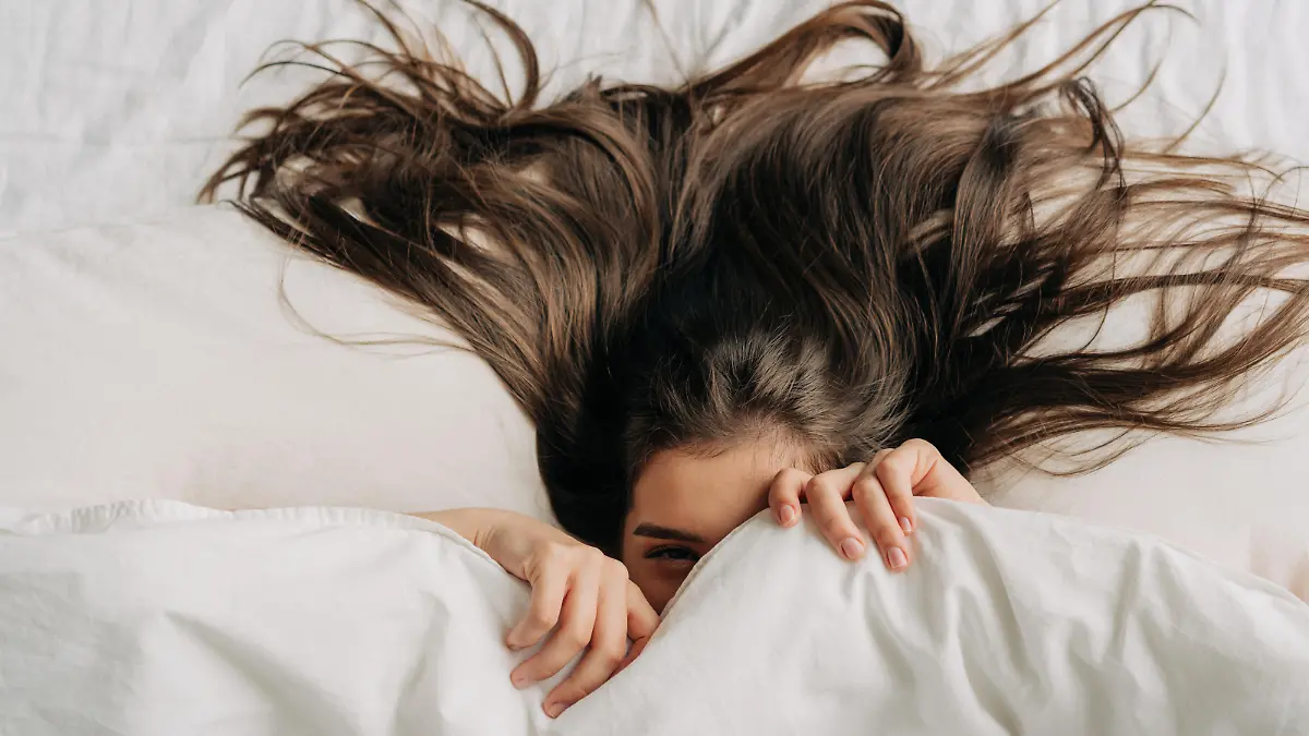 Young funny brunette woman is hiding under the covers while relaxing in bed.