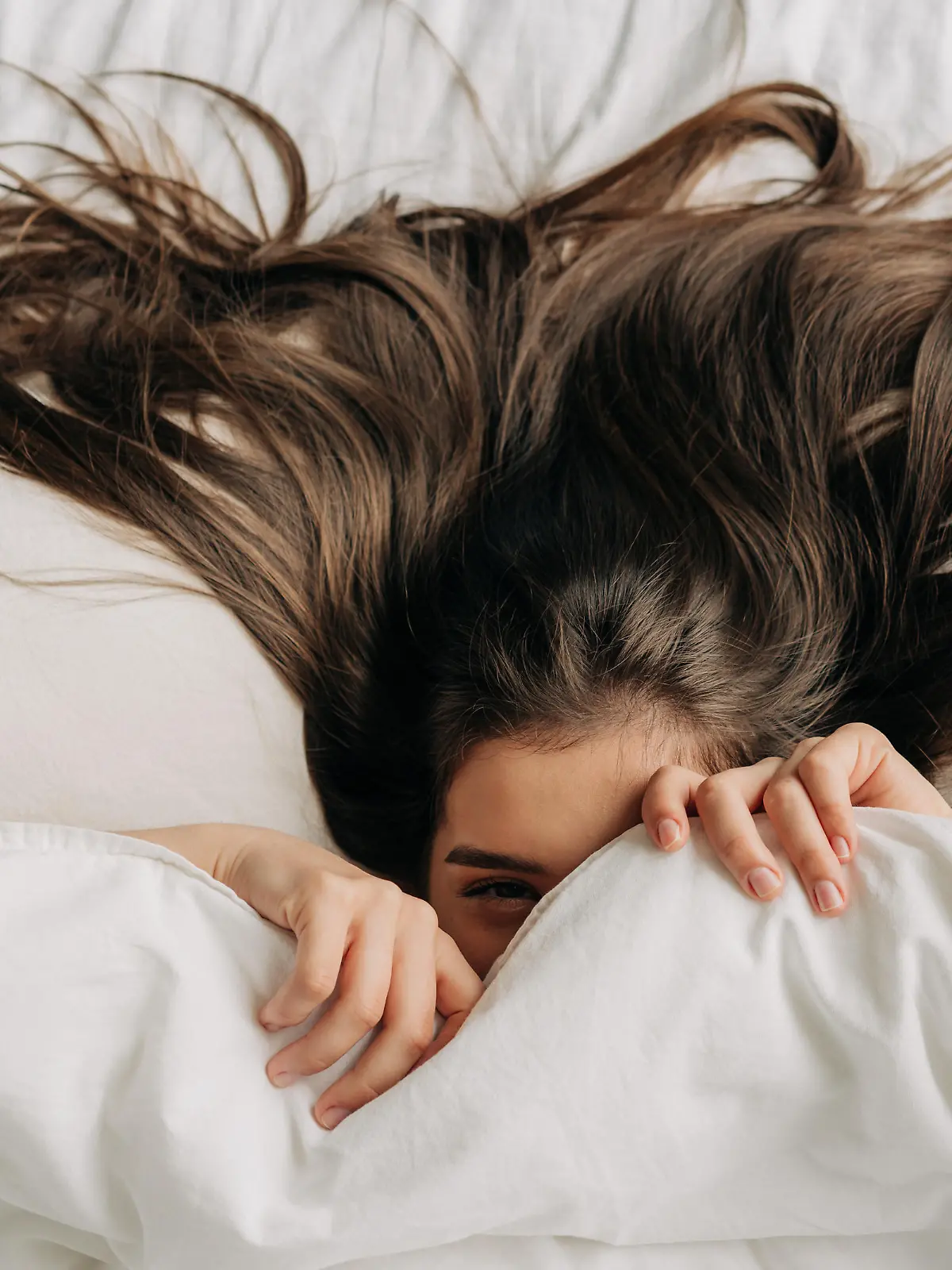 Young funny brunette woman is hiding under the covers while relaxing in bed.