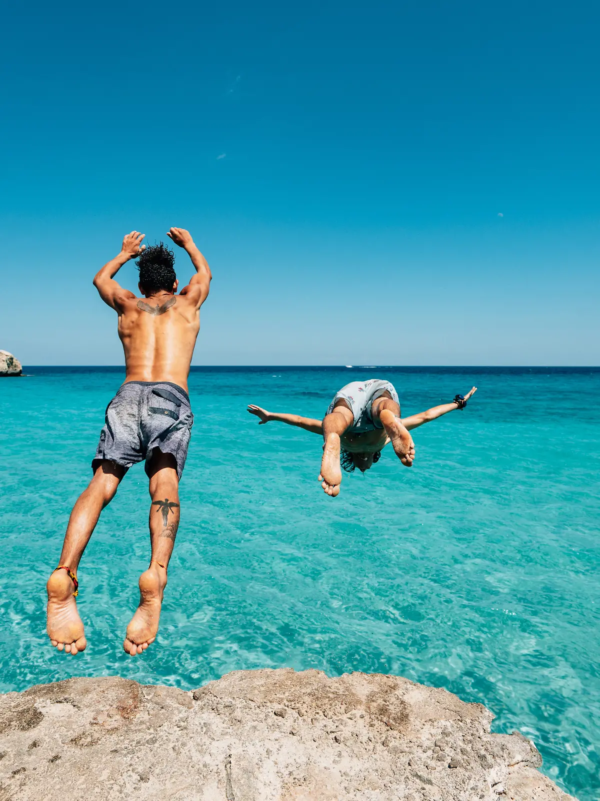 Zwei Freunde springen von einer Klippe ins blaue Mittelmeer.