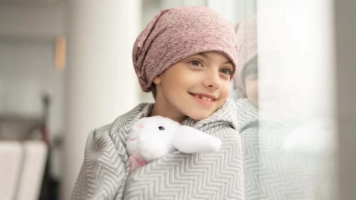 A happy elementary age girl with cancer is wearing a pink scarf on her head. She has a blanket draped over her shoulders and is holding a stuffed toy. The child is smiling boldly while leaning against a window.