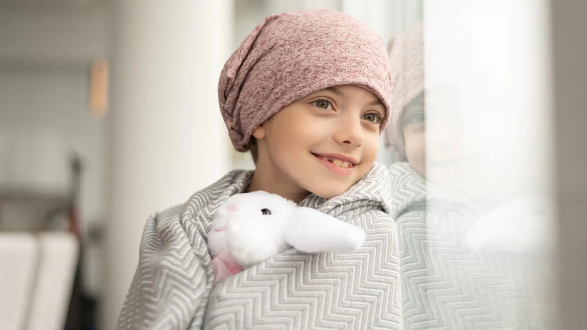 A happy elementary age girl with cancer is wearing a pink scarf on her head. She has a blanket draped over her shoulders and is holding a stuffed toy. The child is smiling boldly while leaning against a window.