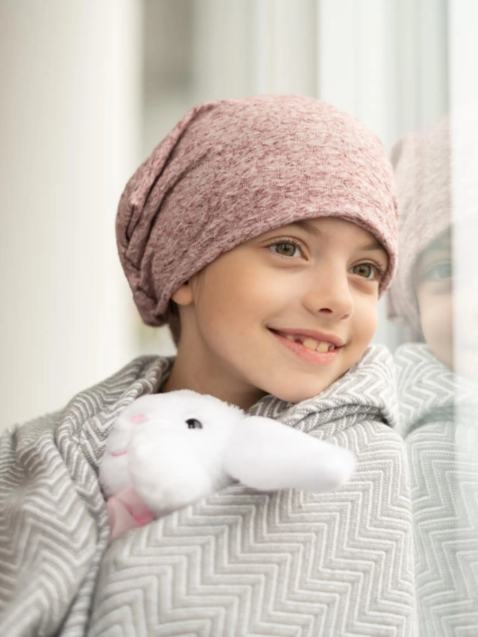 A happy elementary age girl with cancer is wearing a pink scarf on her head. She has a blanket draped over her shoulders and is holding a stuffed toy. The child is smiling boldly while leaning against a window.