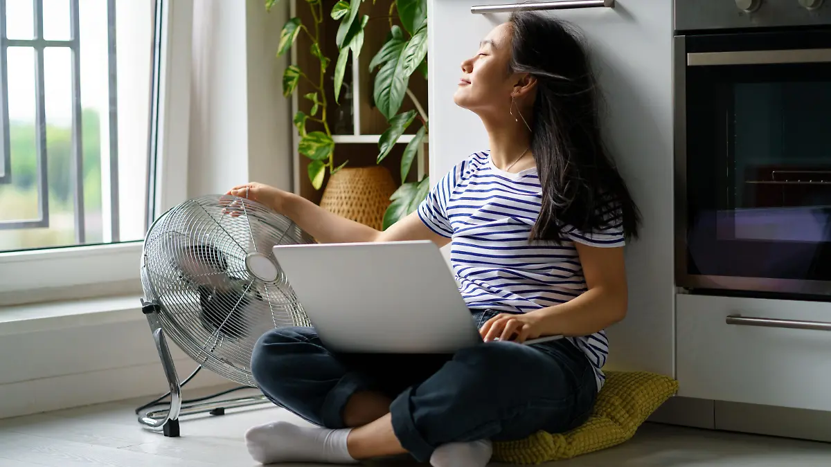 Eine Frau sitzt mit einem Laptop und einem Ventilator auf dem Küchenboden.