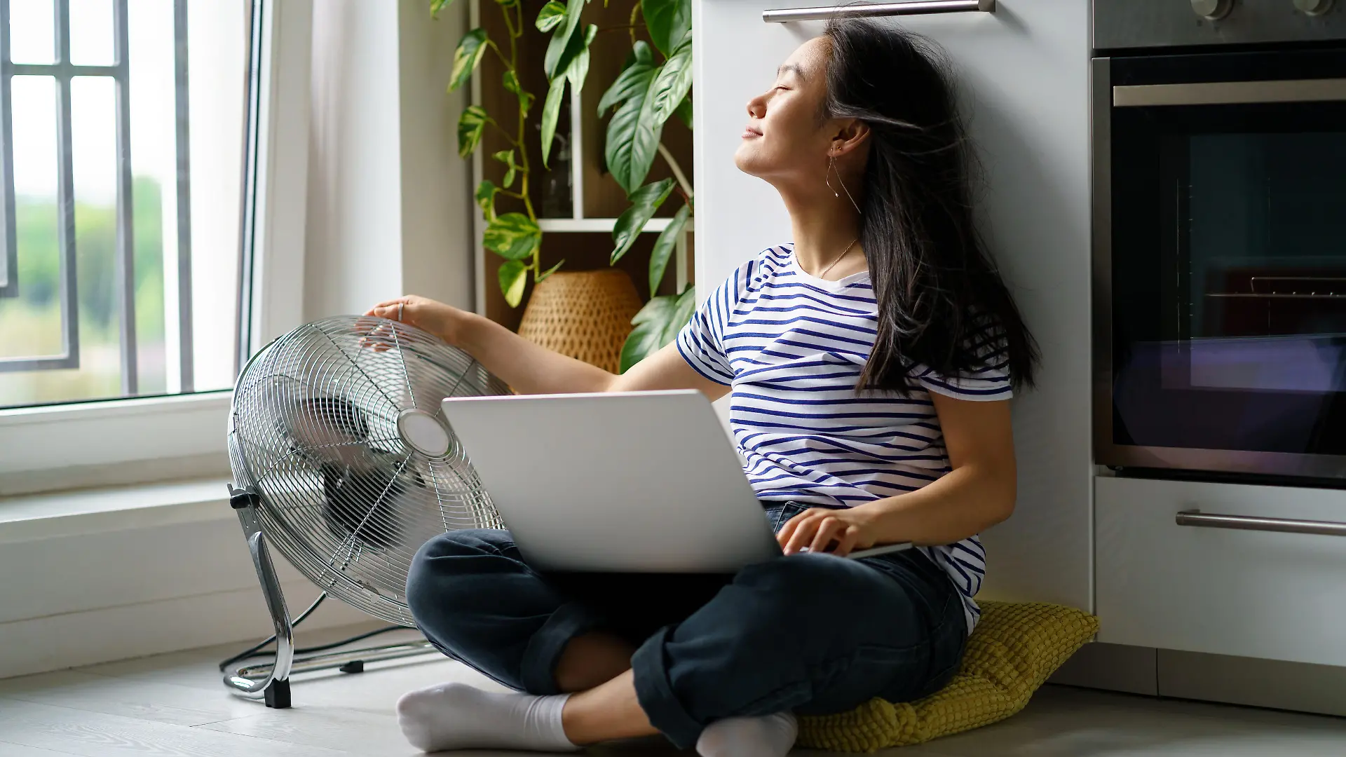 Eine Frau sitzt mit einem Laptop und einem Ventilator auf dem Küchenboden.