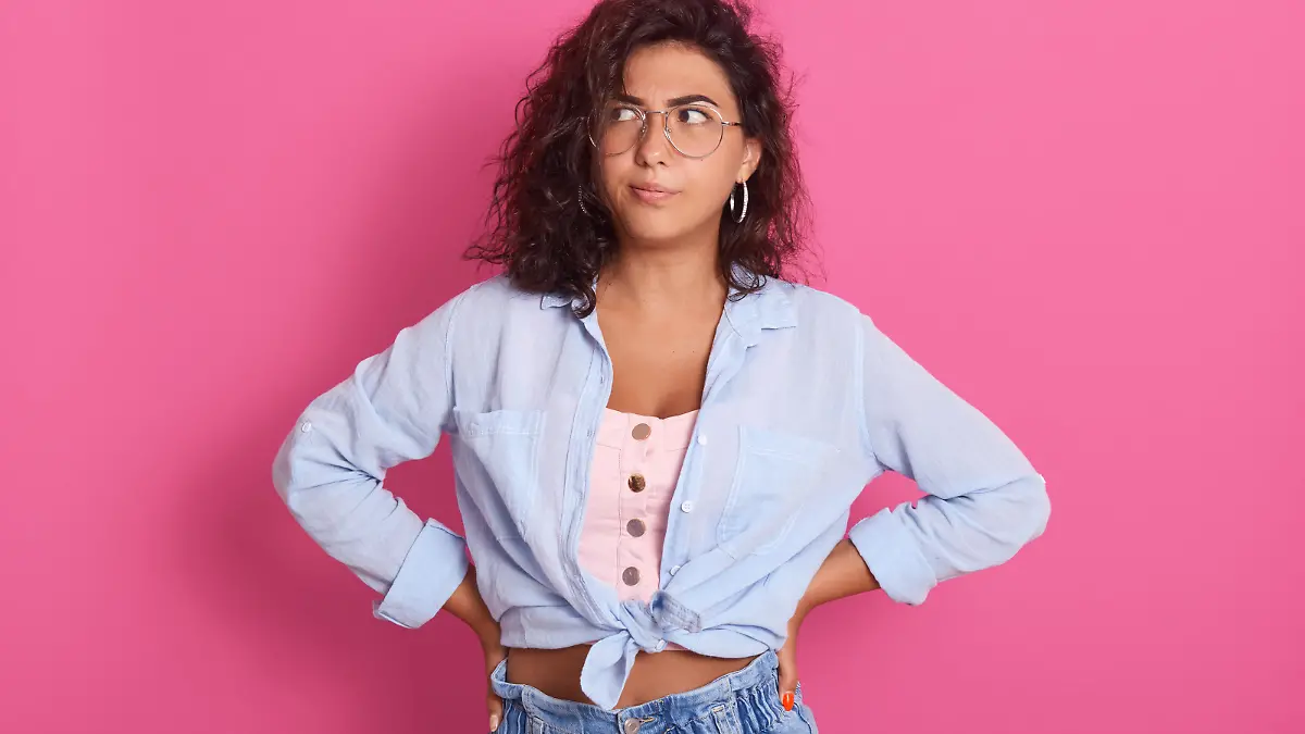 Horizontal shot of young female with thoughtful expression, looks sideways, keeps hands on hips, thinks about something, attractive woman with dark wavy hair poses against pink studio background