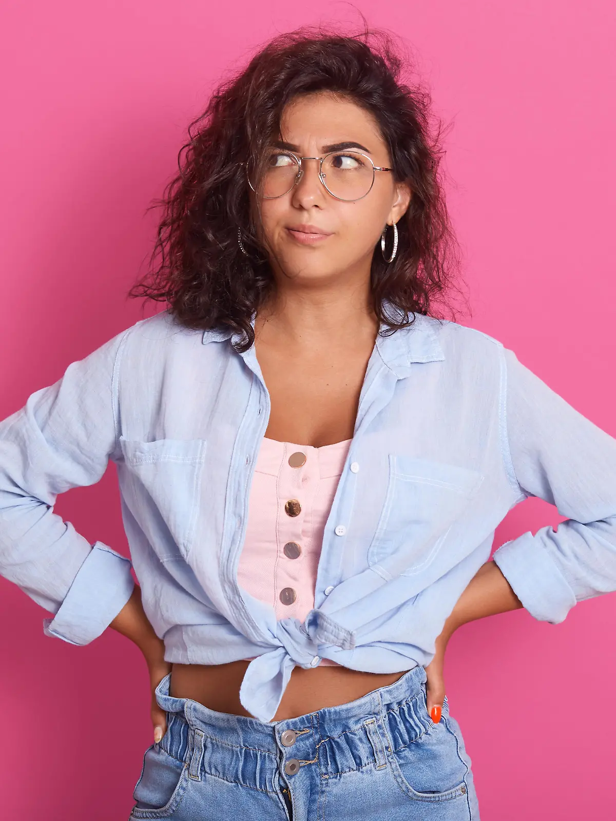 Horizontal shot of young female with thoughtful expression, looks sideways, keeps hands on hips, thinks about something, attractive woman with dark wavy hair poses against pink studio background