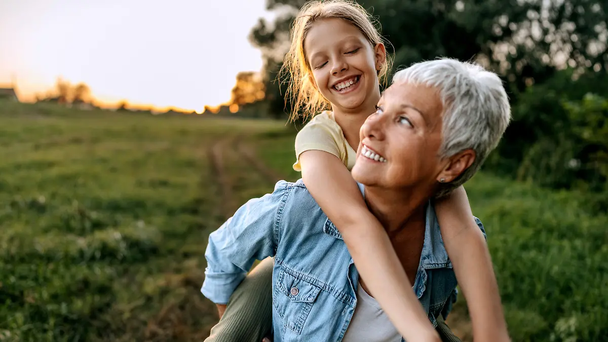 Oma trägt ihr Enkelin huckepack über eine Wiese, beide lächeln glücklich