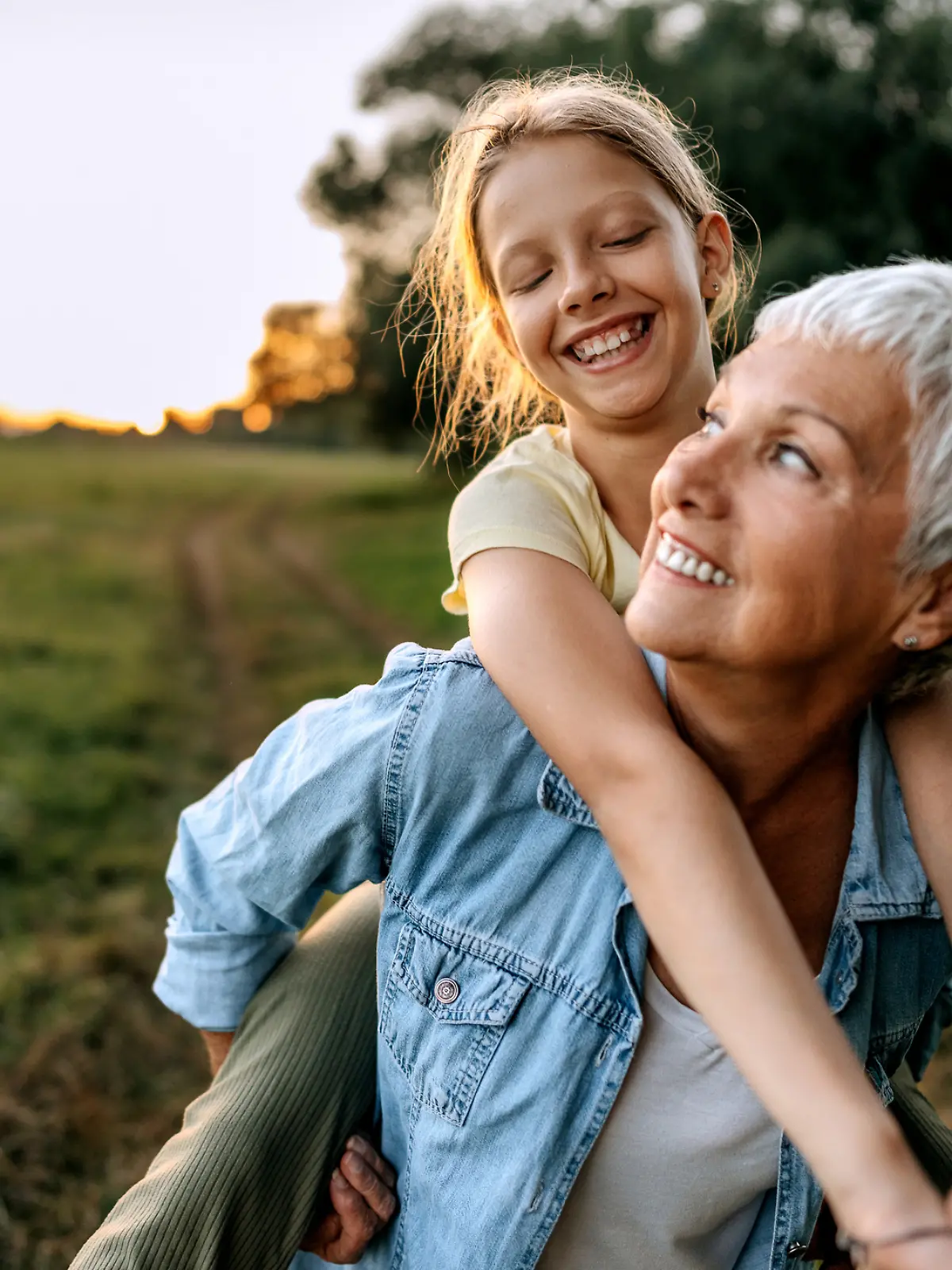 Oma trägt ihr Enkelin huckepack über eine Wiese, beide lächeln glücklich