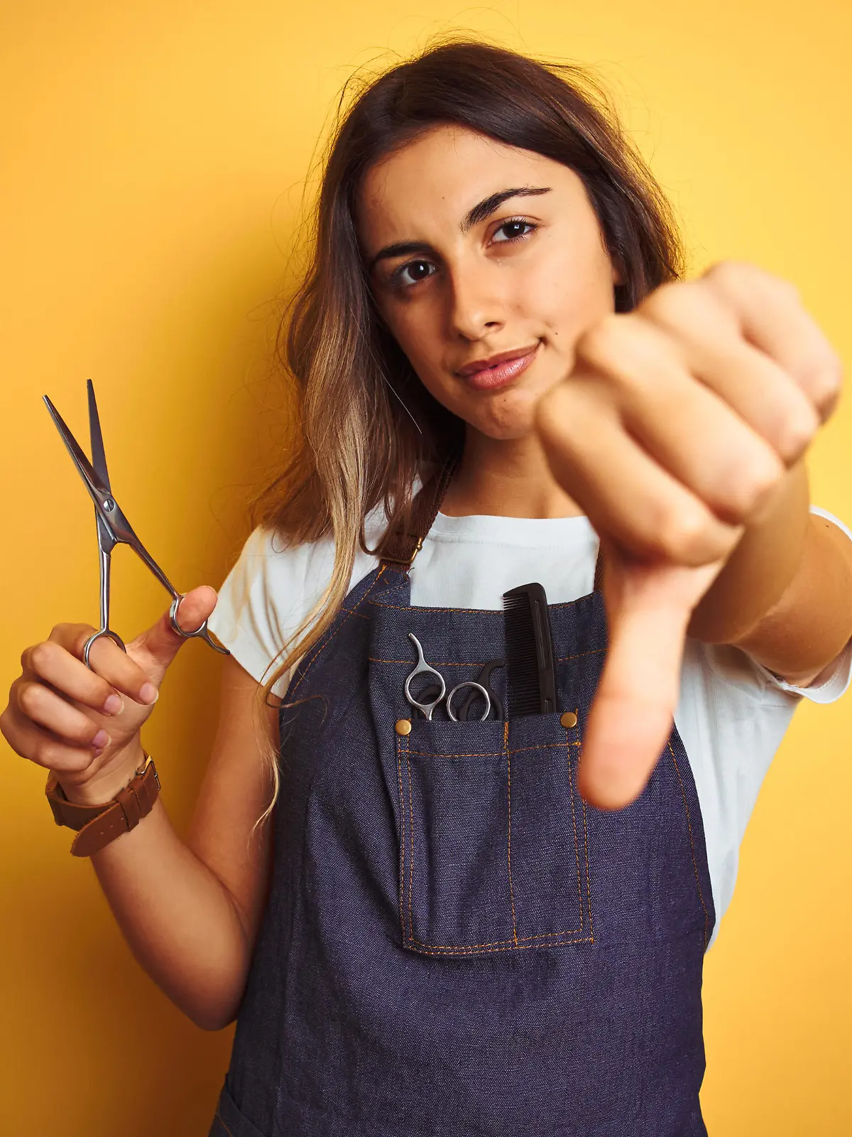 Young beautiful hairdresser woman holding scissors over yellow isolated background with angry face, negative sign showing dislike with thumbs down, rejection concept