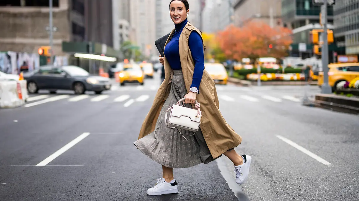 Beautiful woman in a beige sleeveless coat, skirt, turtleneck sweater, white sneakers and with a handbag seen crossing a street in Manhattan, New York, while using her wireless headphones and talking on the phone during a break at work.