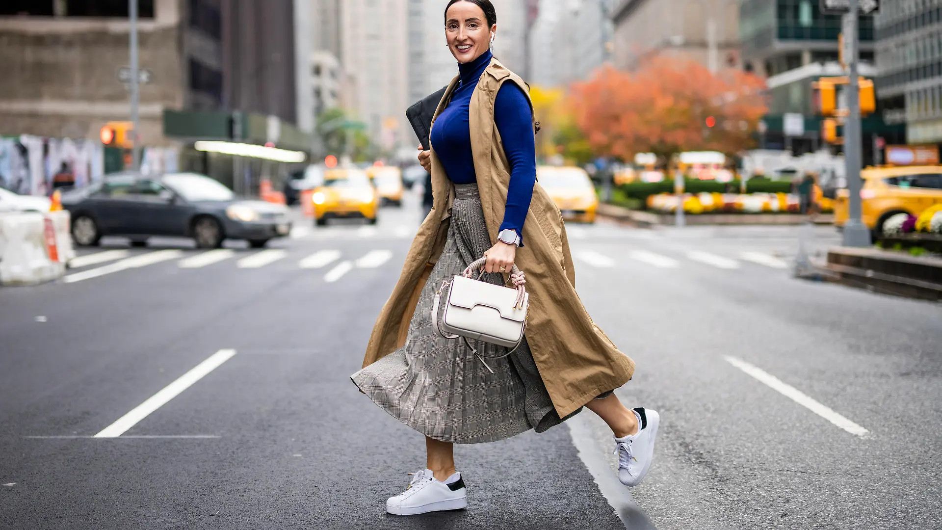 Beautiful woman in a beige sleeveless coat, skirt, turtleneck sweater, white sneakers and with a handbag seen crossing a street in Manhattan, New York, while using her wireless headphones and talking on the phone during a break at work.