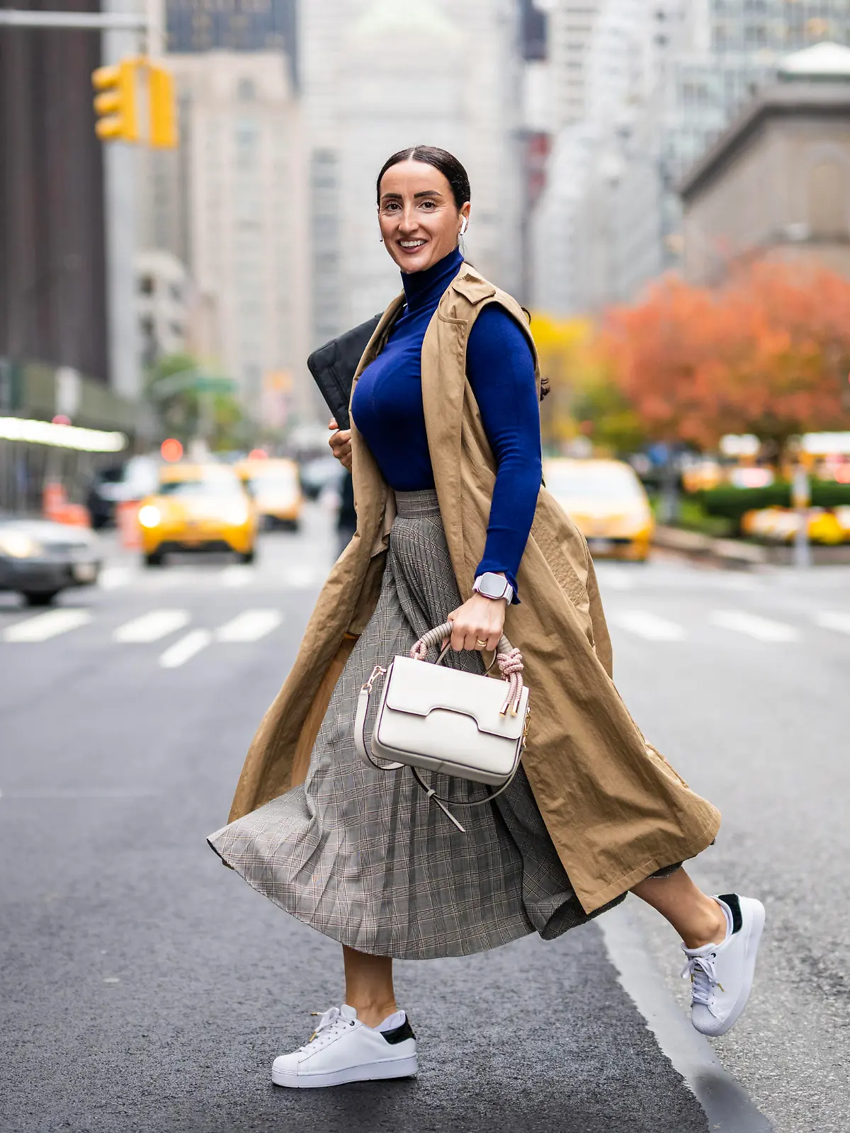 Beautiful woman in a beige sleeveless coat, skirt, turtleneck sweater, white sneakers and with a handbag seen crossing a street in Manhattan, New York, while using her wireless headphones and talking on the phone during a break at work.