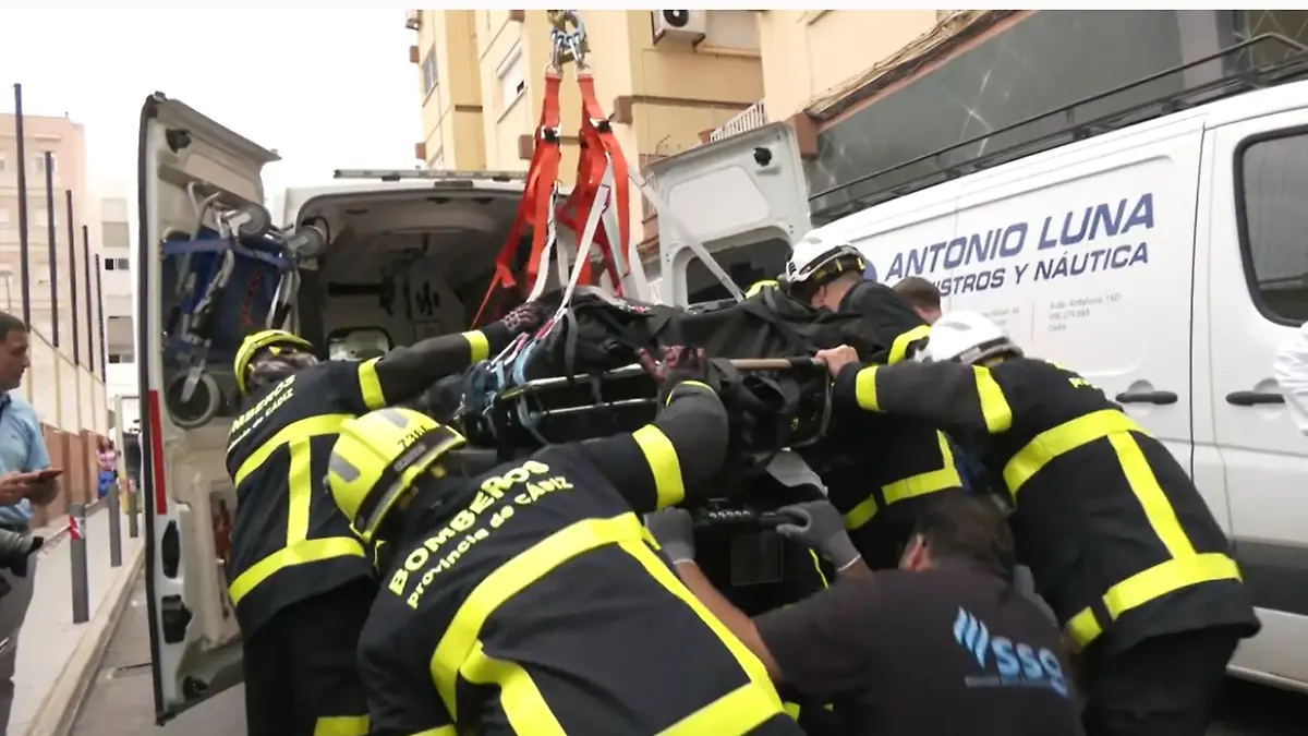 Feuerwehrleute laden einen sehr schweren Mann auf einer Trage mit einem Kran in einen Krankenwagen.