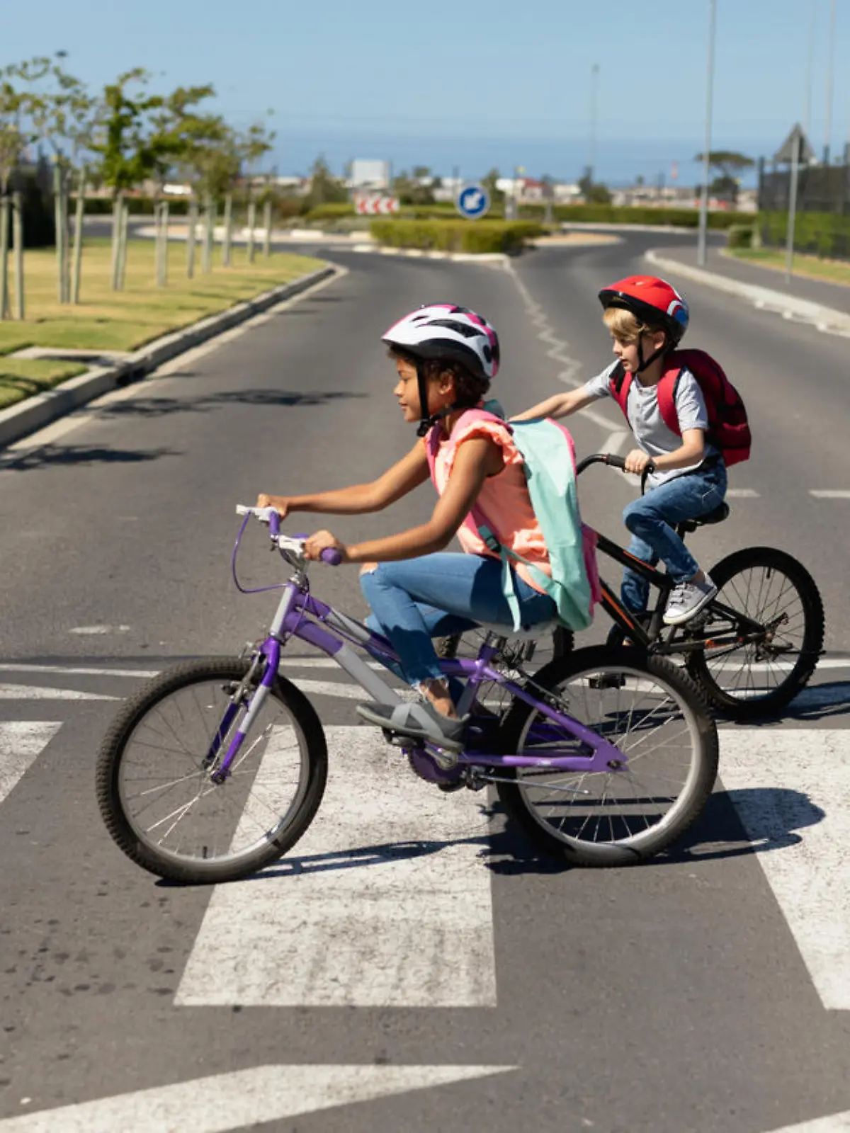 Side view of a Caucasian and an African American schoolgirl, both wearing cycling helmets and riding bicycles, crossing the road on a pedestrian crossing, on their way to elementary school on a sunny day