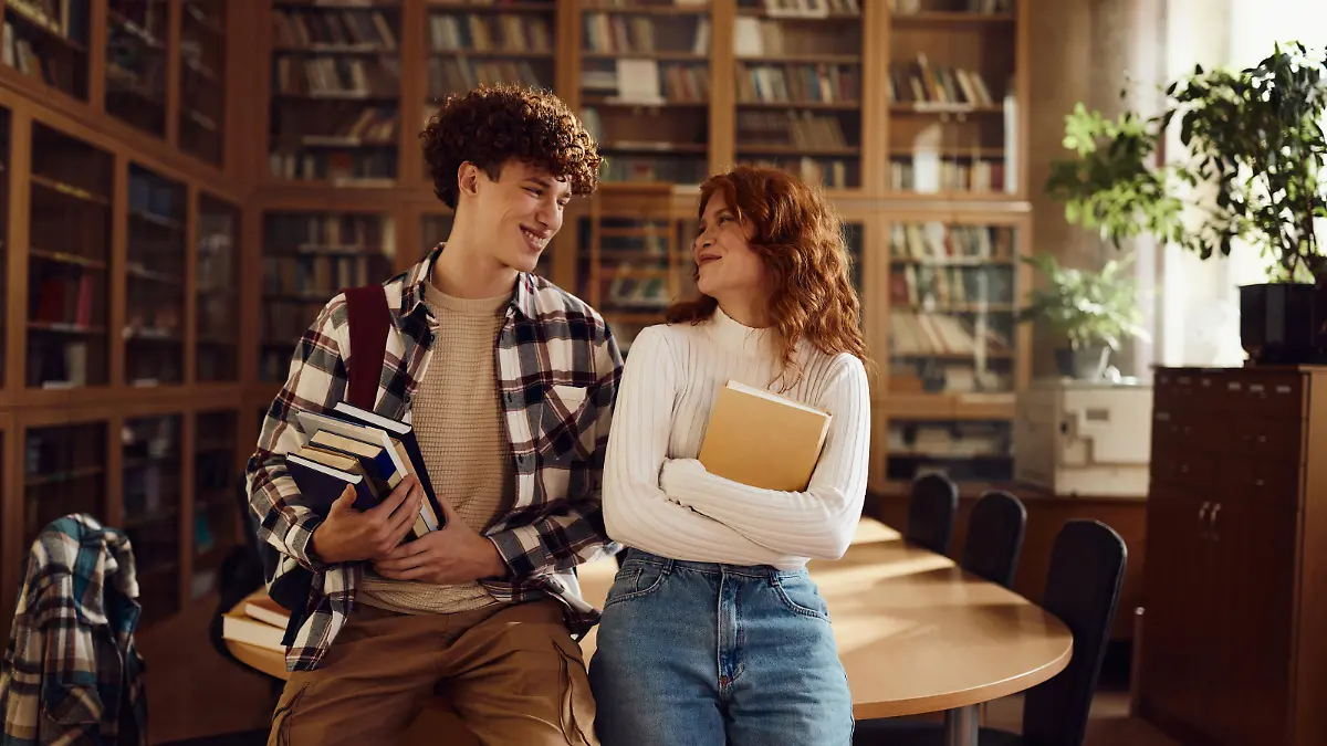 Junger Mann und junge Frau lächeln sich an in einer Bibliothek.