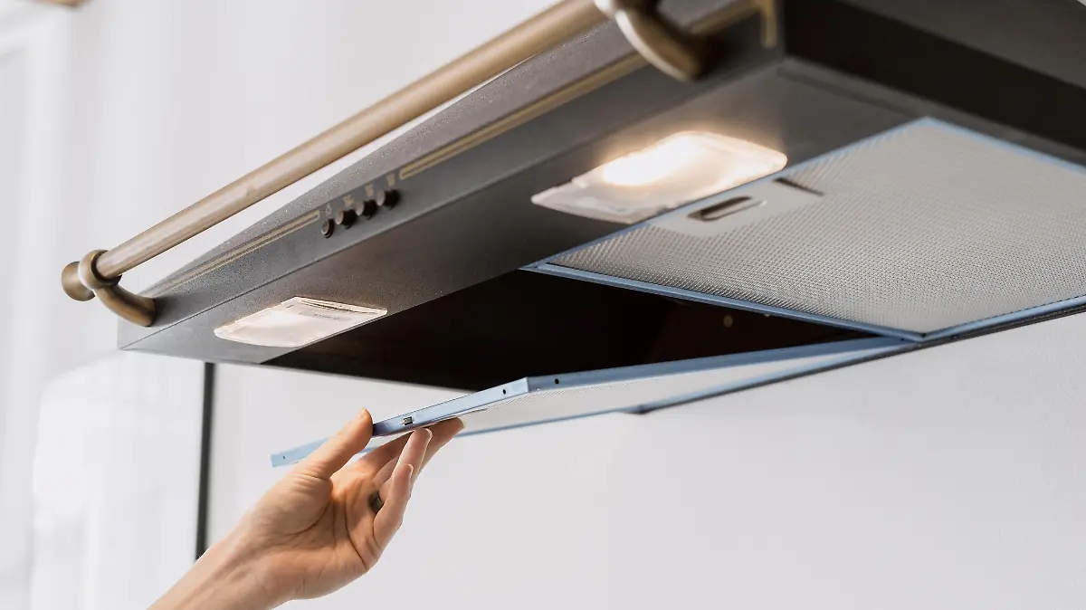 cropped shot of black metal hood extractor and person hand taking aluminum mesh filter on white wall background, maintenance household appliances
